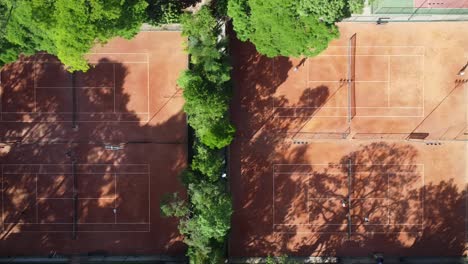 traveling-to-the-side-with-drone-filming-down-beautiful-tennis-courts-in-the-middle-of-trees-and-nature,-sunrise,-soft-light