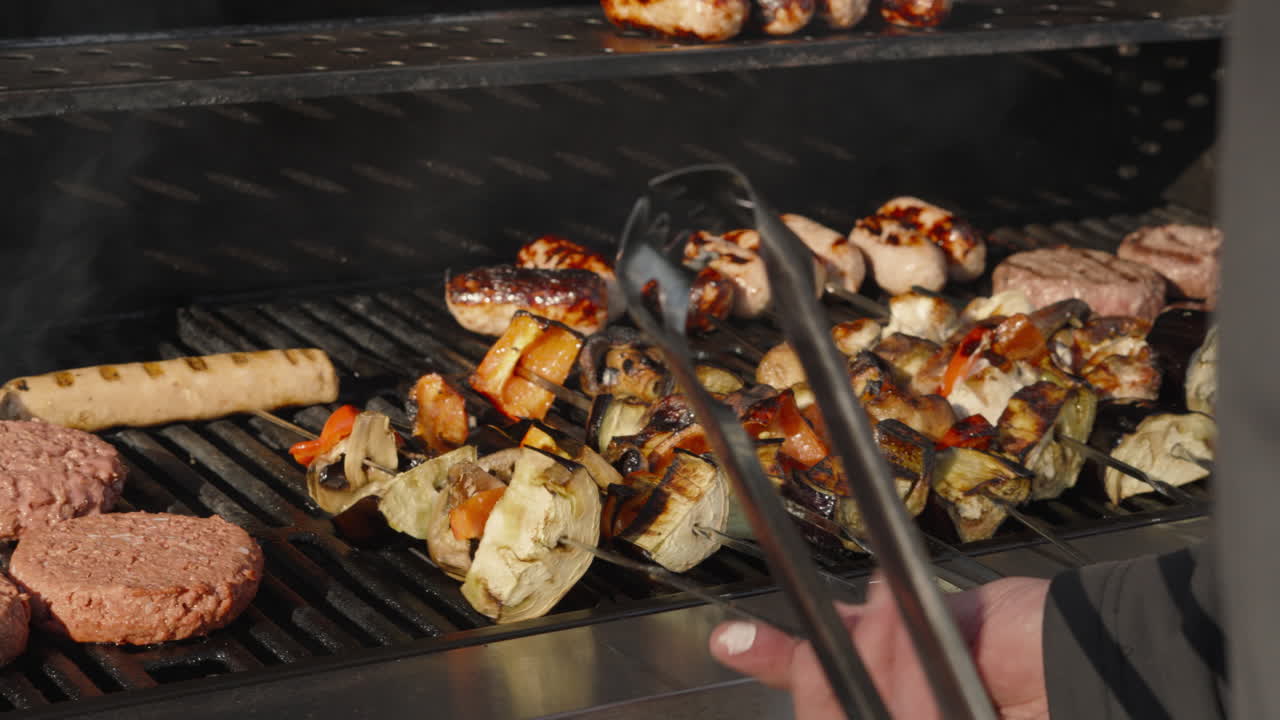Mano dello chef che gira spiedini di verdure sul barbecue con hamburger di manzo e salsicce