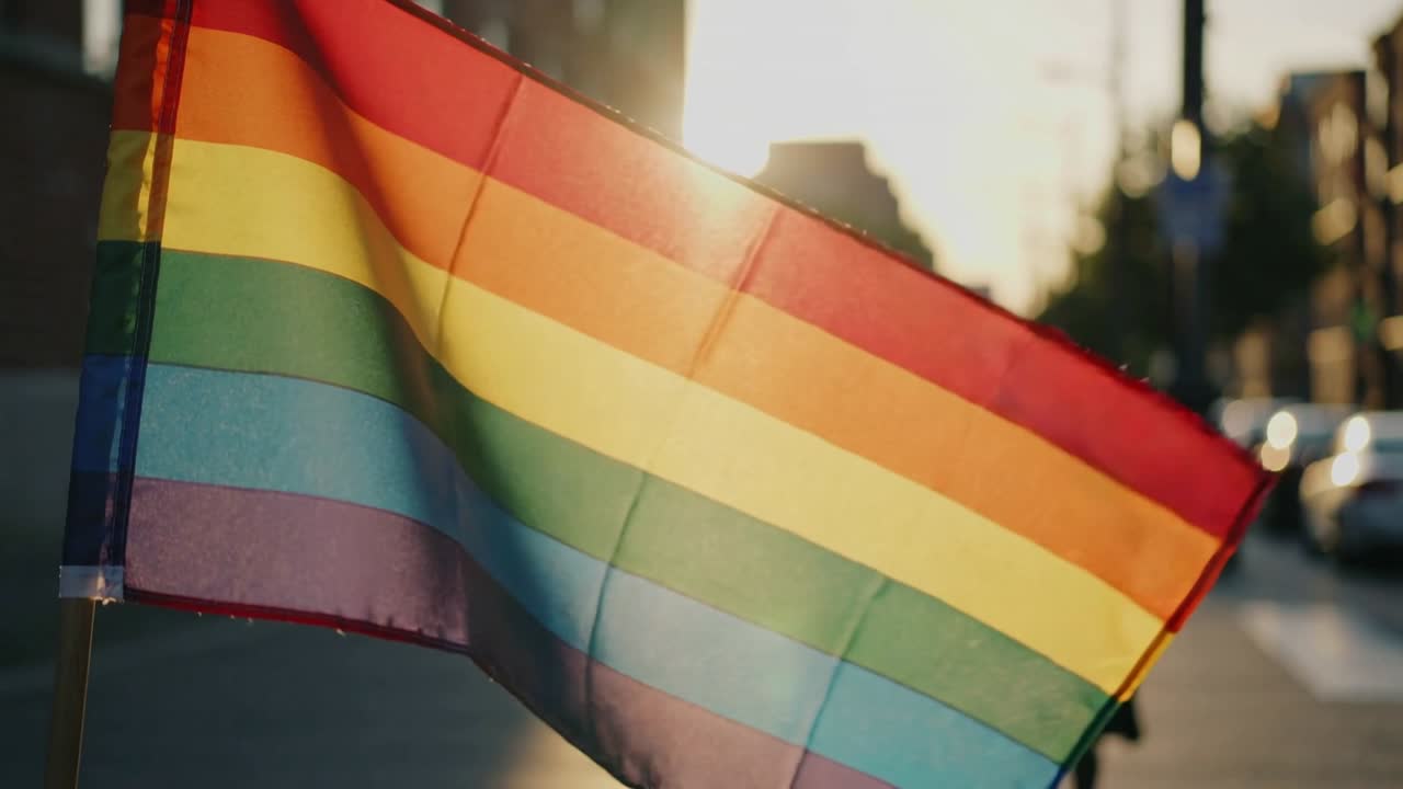 Marketing scene: rainbow pride flag fluttering on pole, breeze causing sun flare and silhouettes