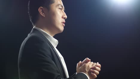 close up side view of asian speaker man in business suit holding his hands together and showing index fingers up while speaking in the black screen studio