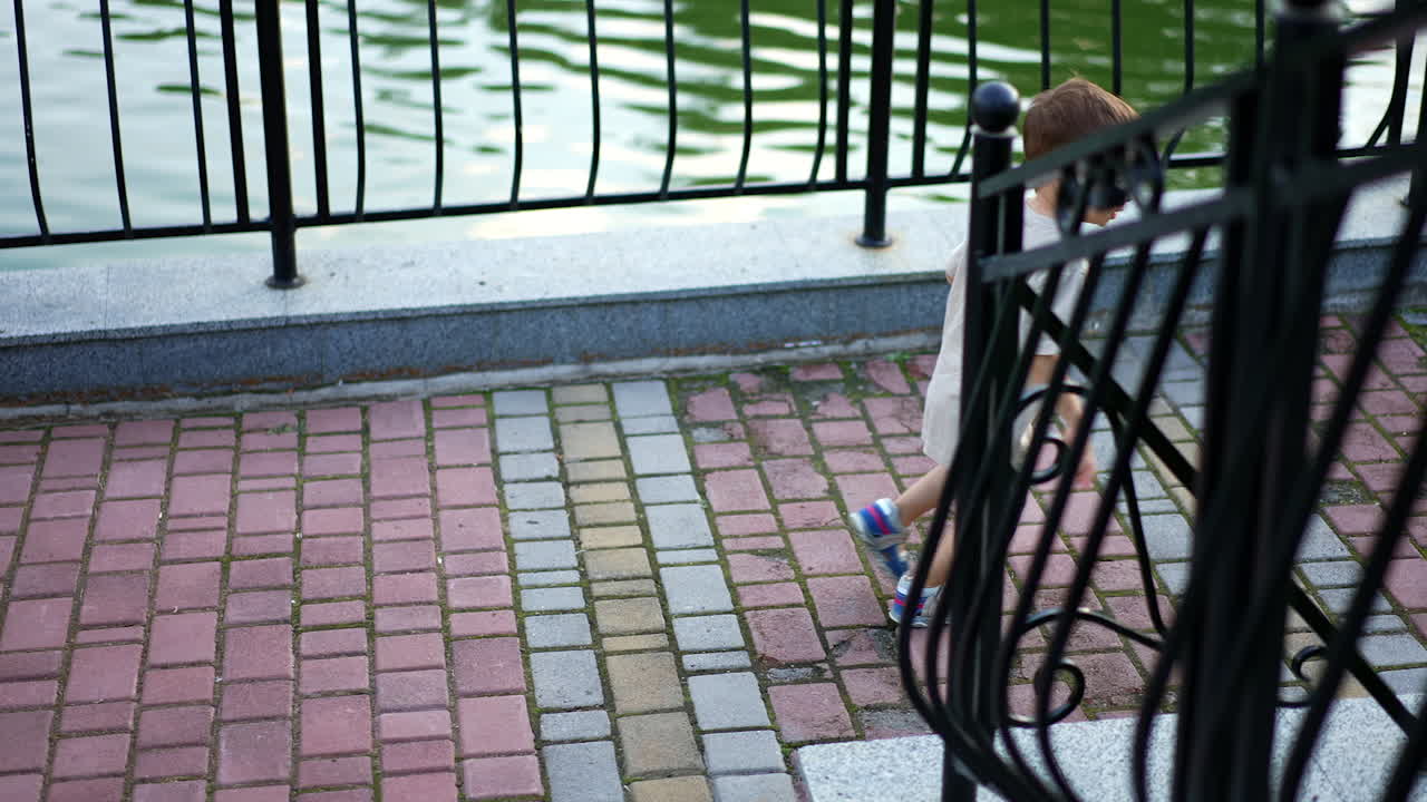 Rear view of baby going downstairs holding by the handrails. Kid runs by the paved alley near the pond.