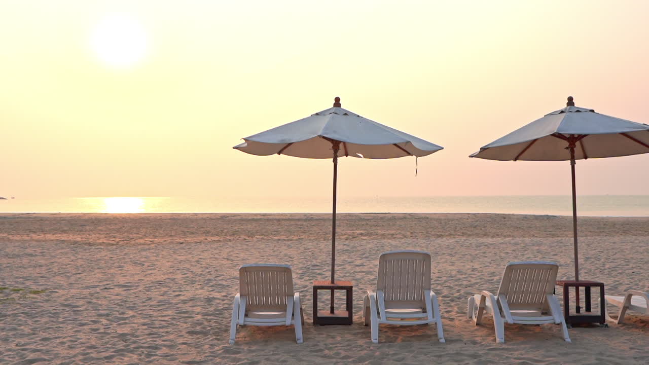 그늘 파라솔 아래 빈 해변 의자는 지는 열대 태양을 바라보며 해변을 가로질러 보입니다.