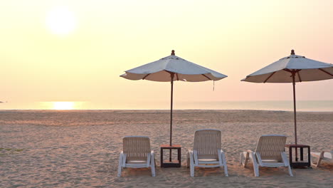 empty beach chairs under shade umbrellas look out across the beach at a setting tropical sun