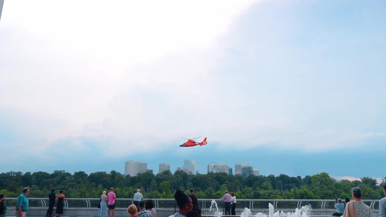 Washington, USA, 13 giugno 2026: Elicottero della guardia costiera rosso in volo sopra il fiume Potomac vicino al Kennedy Center. Velivolo arancione incorniciato da una colonna di marmo contro un cielo nuvoloso con lo skyline di Rosslyn sullo sfondo.