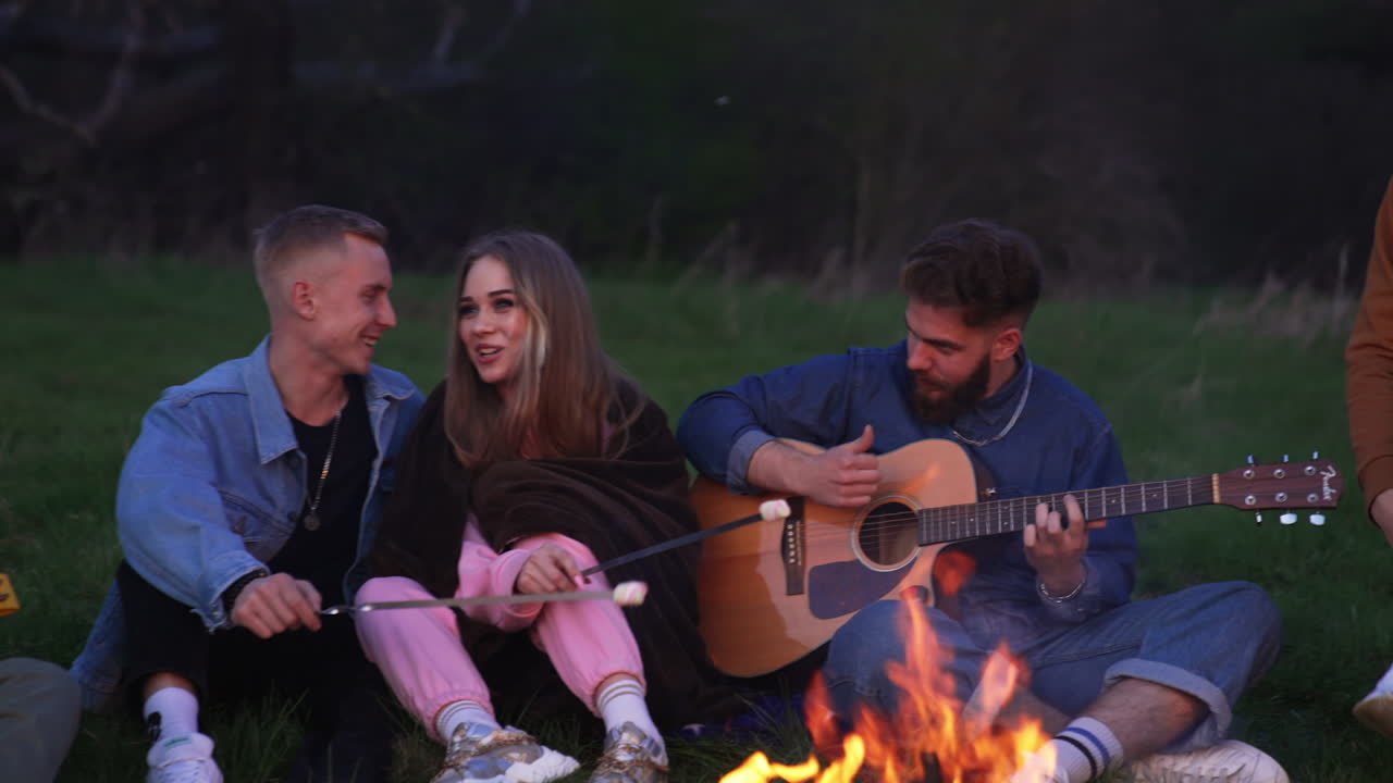 Cheerful youngsters enjoying summer holidays. Playing guitar, singing and frying marshmallows by the campfire in the nature.