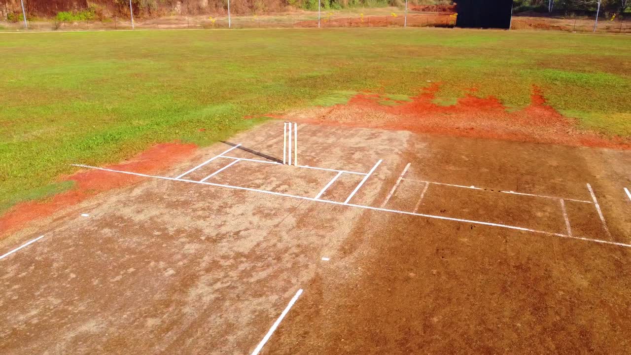 Ripresa aerea con drone di un campo da cricket professionale con linee bianche su terra rossa, circondato da un lussureggiante campo esterno in erba verde in pieno giorno.