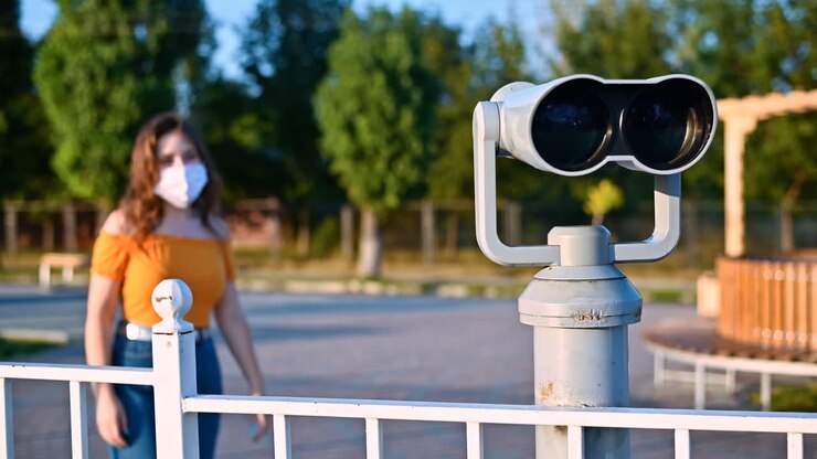 Young woman in protective medical mask looking into a tower viewer from the observation deck at the city at sunset. Corona Virus idea. Safe travel during the pandemic