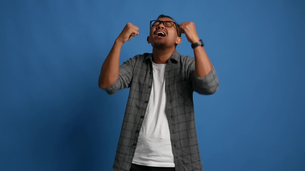Hombre adulto asiático feliz celebrando el éxito mientras levanta los puños, aislado sobre fondo azul