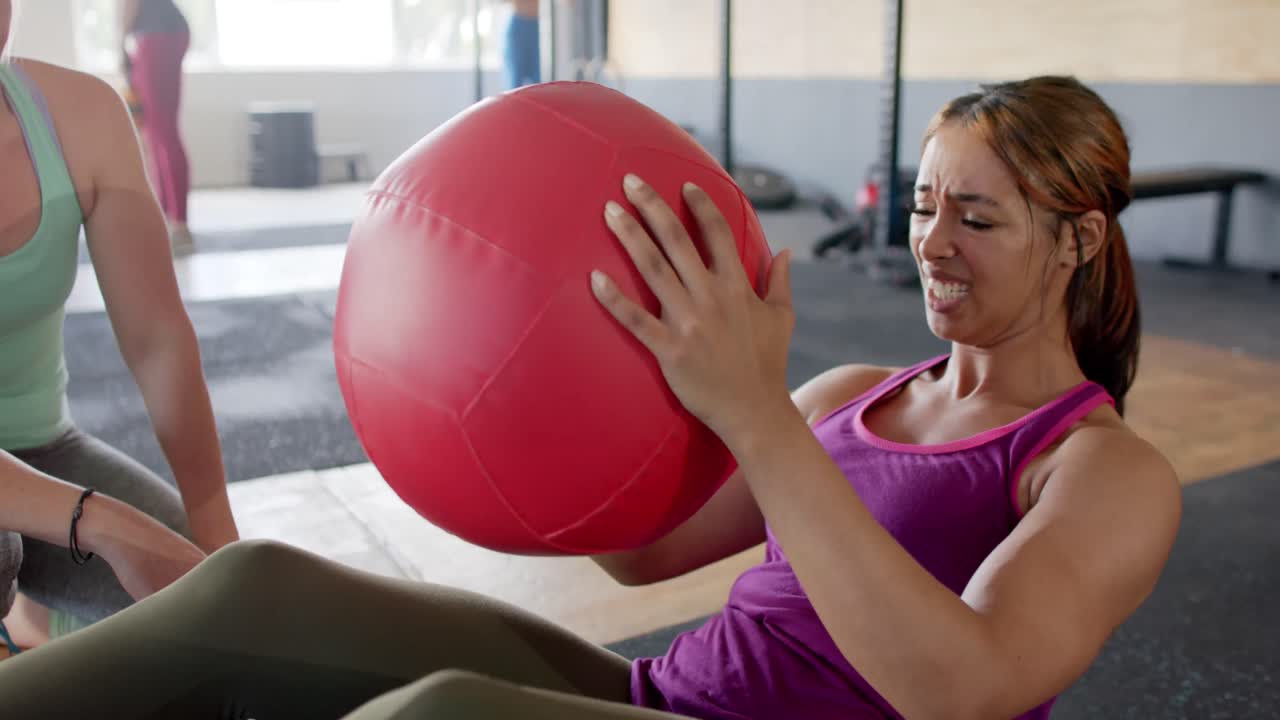 mujer diversa inalterada abrazando a una mujer agotada entrenando con una pelota de medicina en el gimnasio, cámara lenta