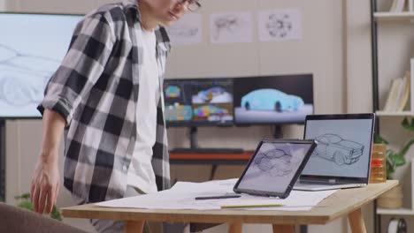 vista lateral de un hombre asiático caminando para sentarse y dibujar un nuevo concepto de diseño de automóvil en una tableta en el estudio con televisión y computadoras muestra modelo de automóvil eléctrico 3d