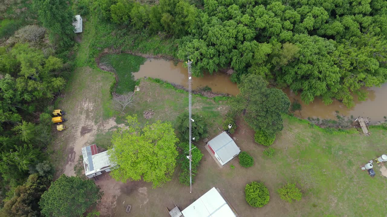 A high-angle, top-down drone view of a remote settlement in the Argentine Delta, featuring a tall communication antenna, solar panels, and simple rural houses