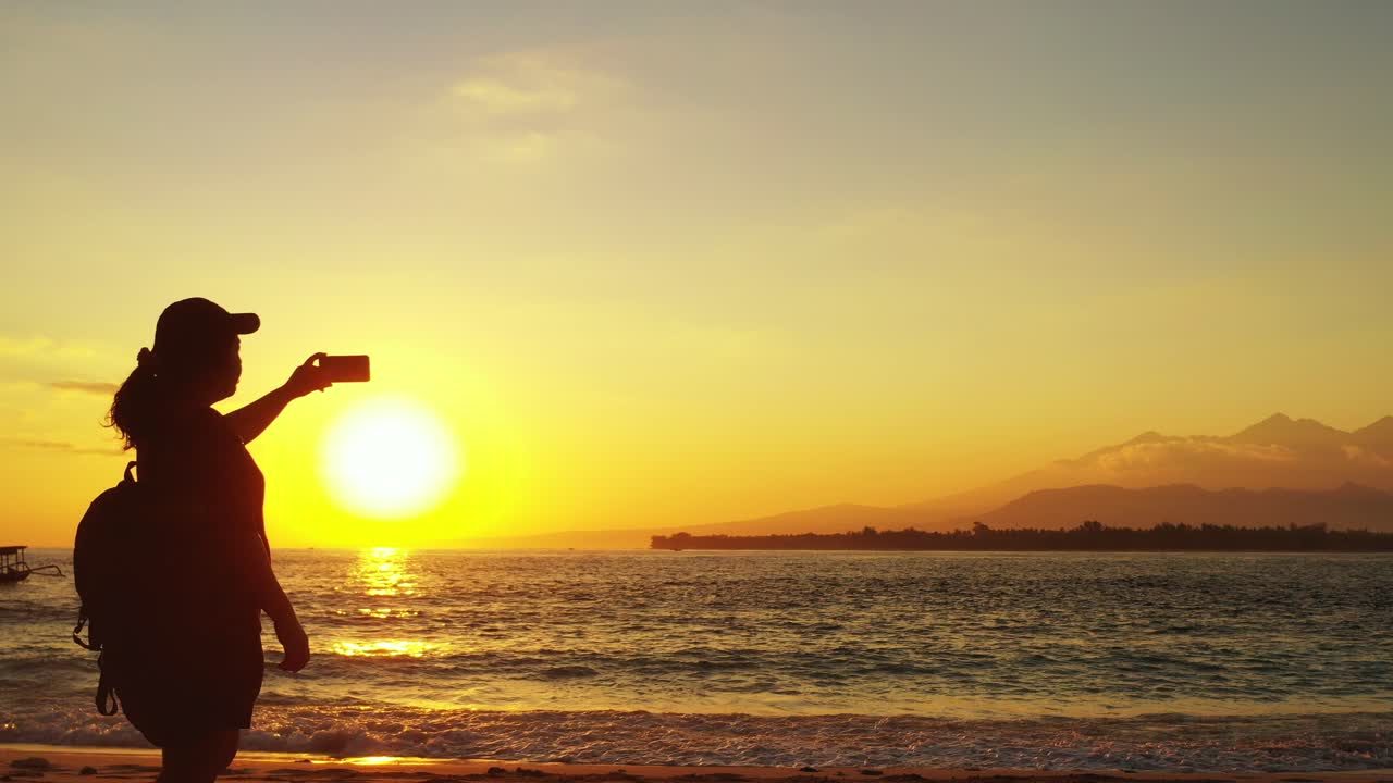 Silhouette of young woman taking photos of beautiful sunset with yellow misty sky reflecting on sea surface near shore of tropical island