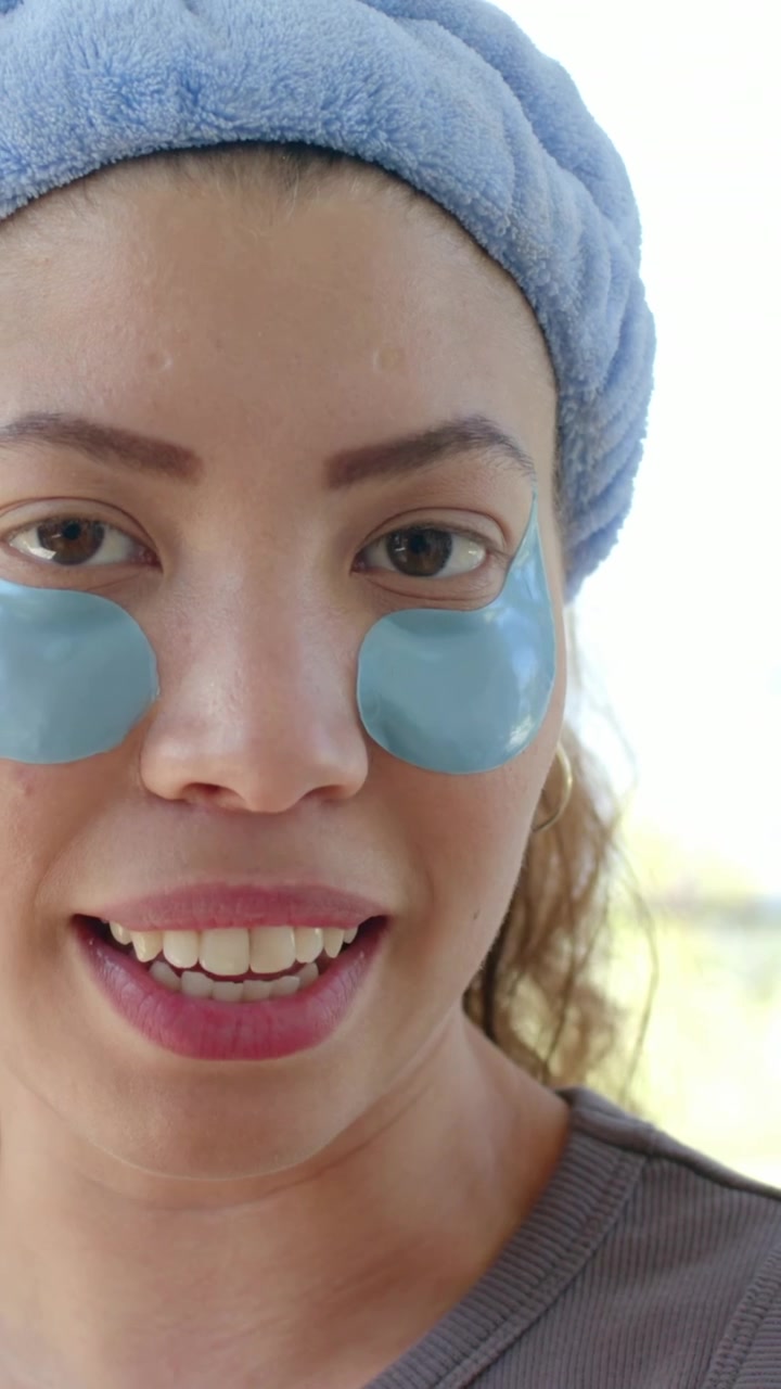Video vertical: Mujer abriendo la boca haciendo su rutina de cuidado de la piel junto a la ventana, llevando una diadema azul y parches para los ojos.