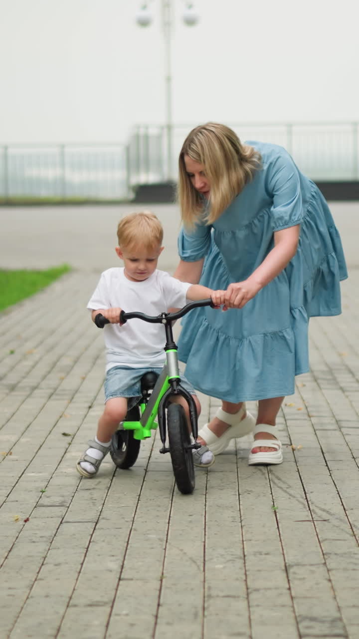 A mother is gently guiding her young son as he rides his bicycle along an interlocked pathway, the boy appears focused, while his mother helps to steady him