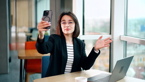 positive caucasian female wearing glasses taking selfie. lady sits at the desk with laptop on. blurred backdrop.