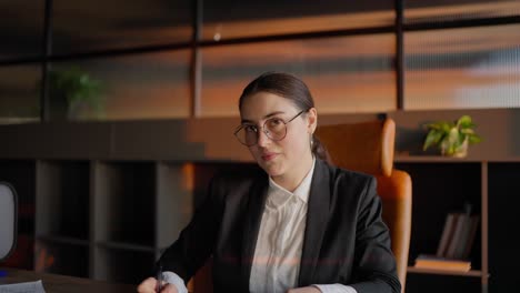 zoom in portrait of a confident concentrated brunette girl in round glasses and a business uniform who sits at a wooden table in the office in front of a laptop and writes down her ideas on paper while working in a sunny office