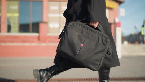 side view of woman carrying black bag with hand adorned with rings, walking confidently, dressed in black attire with blurred red building and decorative greenery in the background