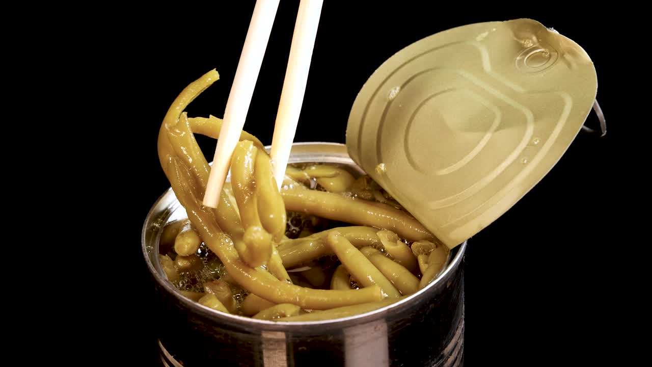 Chopsticks grasp and lift haricot beans from an open tin can against a black background, with steady lighting and a close-up perspective