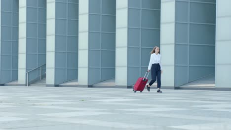 skinny girl in elegant clothes, walking with red suitcase in front of modern building