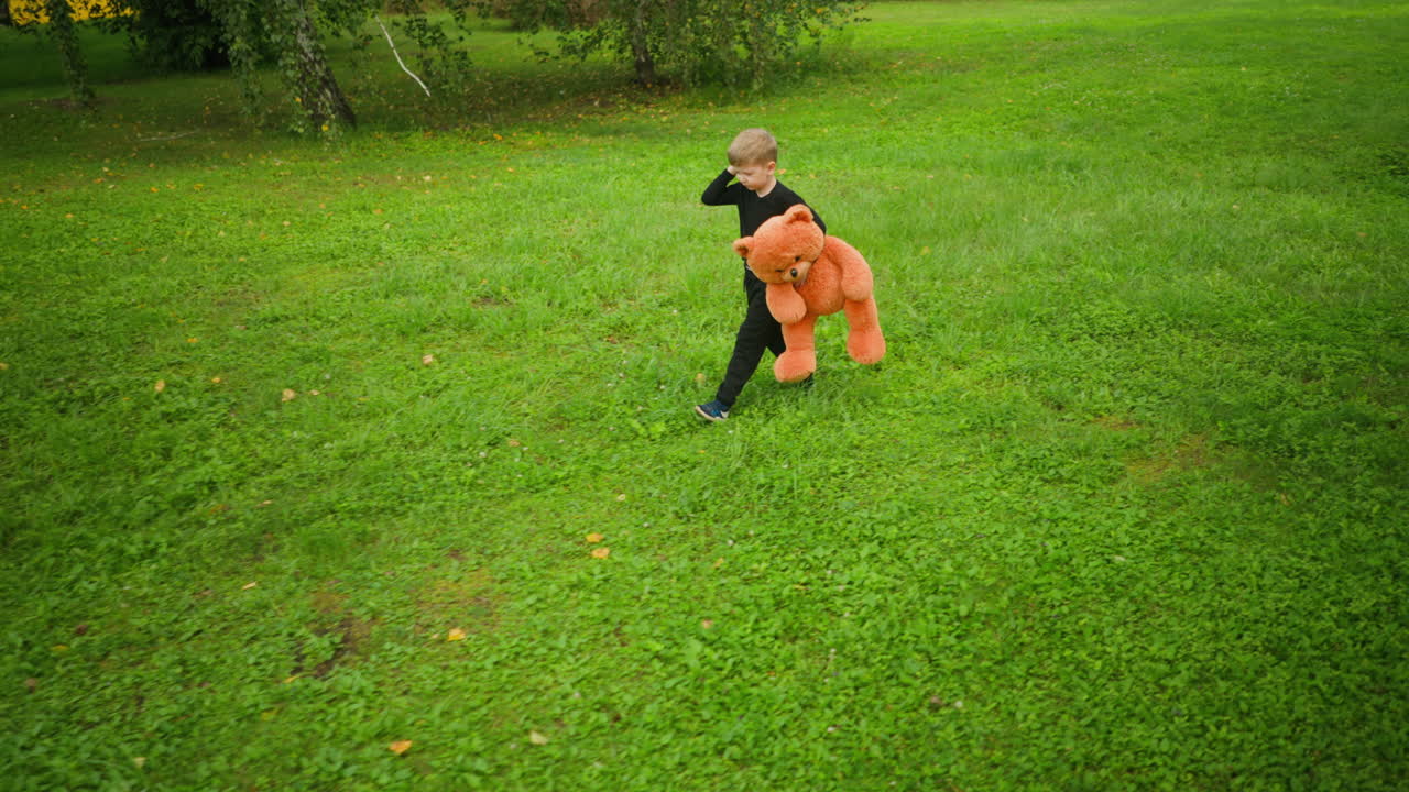 Boy wearing black outfit holding large orange teddy bear while walking slowly across bright green grassy field, raising hand to scratch head thoughtfully, surrounded by trees in quiet environment