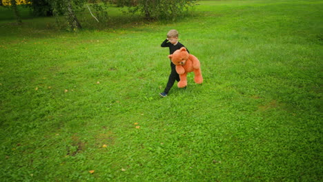 boy wearing black outfit holding large orange teddy bear while walking slowly across bright green grassy field, raising hand to scratch head thoughtfully, surrounded by trees in quiet environment