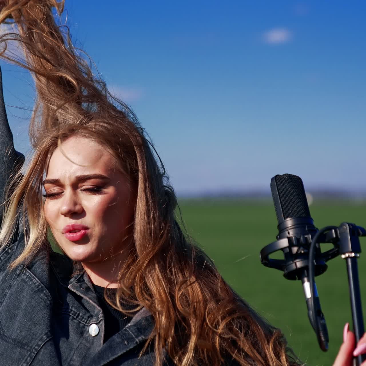 Attractive woman performing on green field background. Beautiful girl in denim suit singing in a microphone and looking at camera outdoors