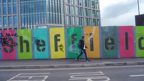 stabilised gimbal panning shot of young man with backpack walking past large sheffield sign, in sheffield city centre, england