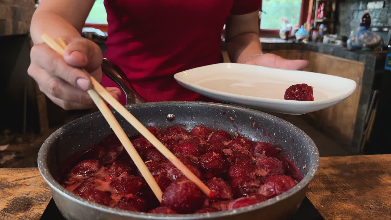 Woman in red shirt serves cooked plums from pot to white plate in cozy kitchen