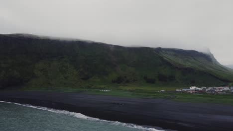 Luftdrohnenaufnahme-Von-Wunderschönen-Blauen-Wellen,-Die-An-Einem-Schwarzen-Sandstrand-In-Island-Zusammenbrechen,-Und-Riesigen-Felsformationen-In-Der-Ferne