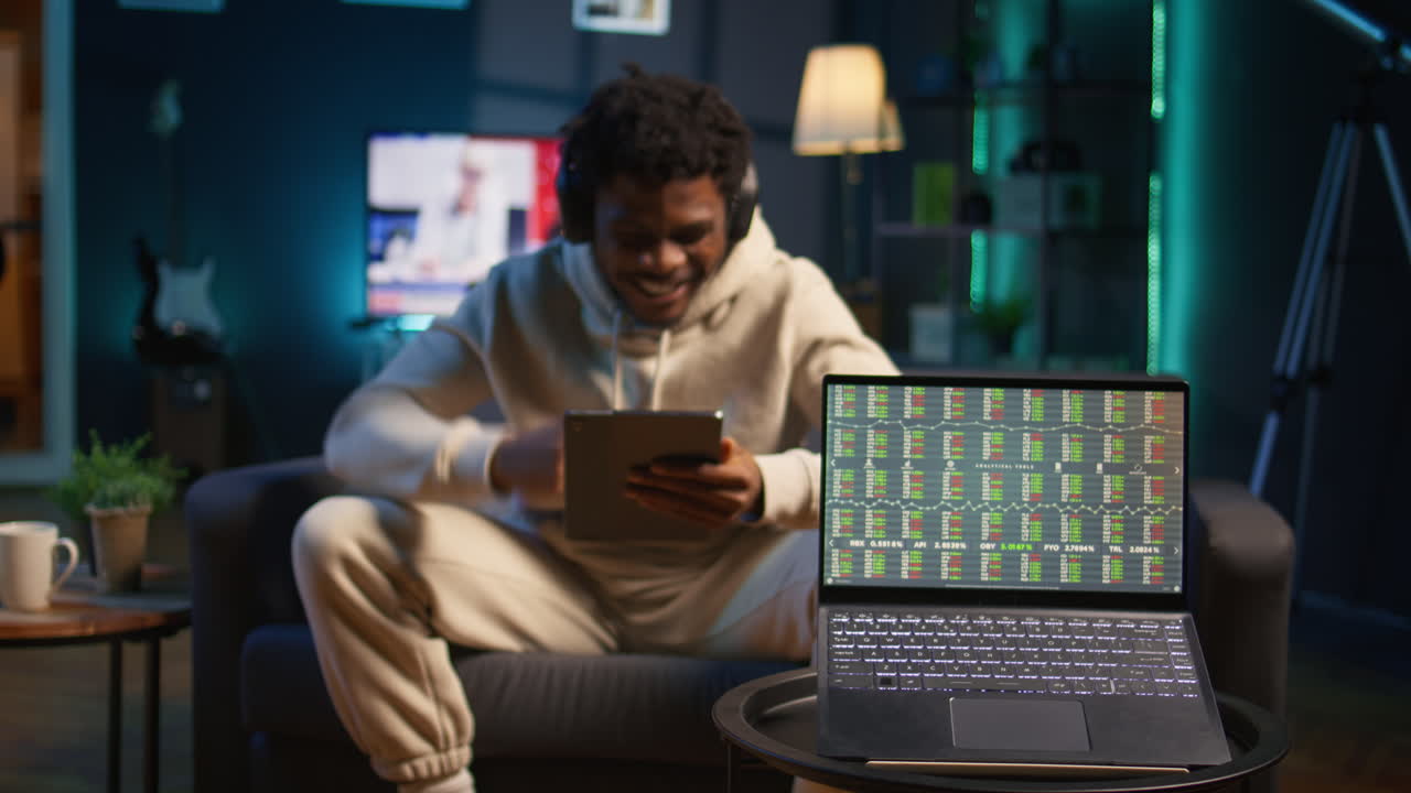 African american person using his tablet laying on the couch while he pays attention to laptop with stock market data trends. Focused man comparing trading graphs numbers before investing. Camera B.