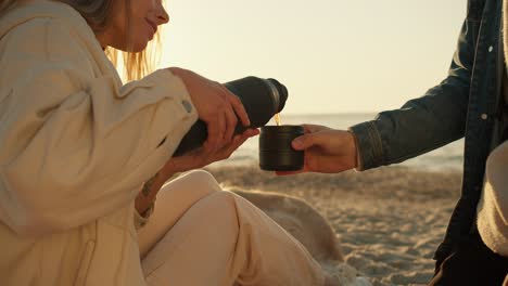 toma de primer plano: una chica vierte té de un termo en un vaso para un chico en una playa soleada por la mañana. sentado en la playa, paseo matutino, atmósfera acogedora