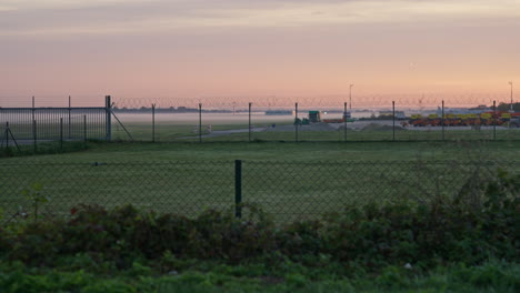 morning light and orange sky behind the airport in munich from outside the fence, with grass in the foreground and fog on the runway