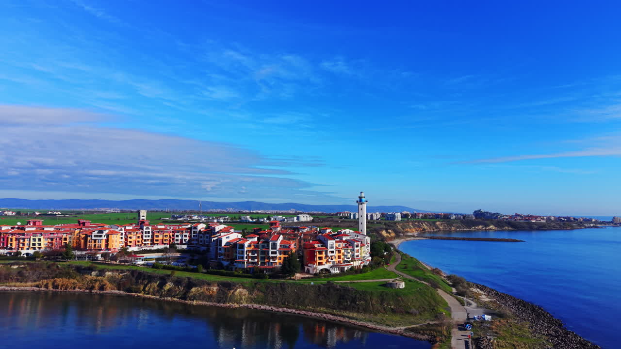 Ripresa dall'alto di una città costiera con condomini colorati, una torre bianca e prati verdi. Zona residenziale costiera con faro e campi verdi, vista aerea.