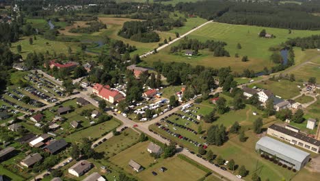 Aerial view of a small town hosting a lively outdoor fair with market stalls, parked cars, houses, and surrounding green fields under a sunny sky. Jaunpiebalga