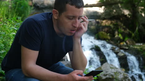 young man holding and smoking a cigarette while browses social media on a smartphone near a waterfall