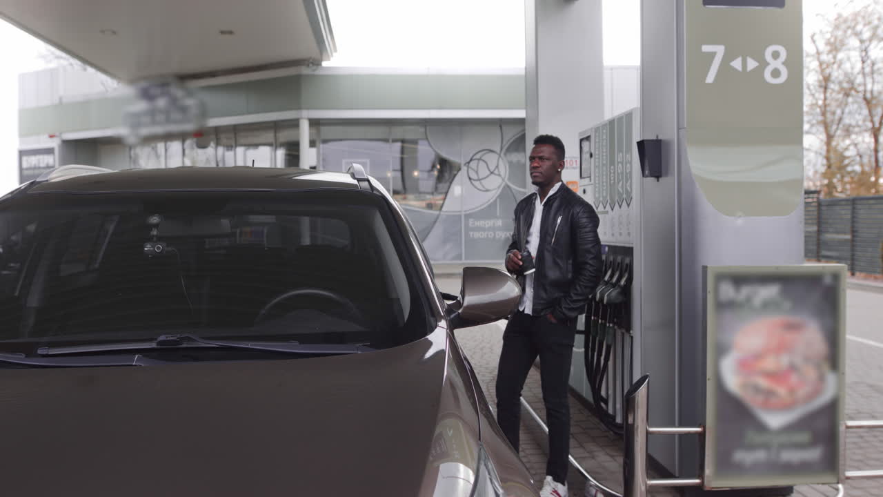 Un giovane e bel uomo d'affari africano in abbigliamento casual nero sta facendo rifornimento alla sua auto moderna a una stazione di servizio