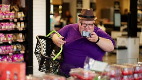 Un-Hombre-Con-Sobrepeso-Con-Una-Camiseta-Morada-Y-Un-Sombrero-Marrón-Huele-Bayas-En-Un-Supermercado-Y-Las-Pone-En-Su-Cesta.-Caminando-Con-Una-Personalidad-Brillante,-Un-Hombre-Con-Sobrepeso-Elige-Bayas-Para-Sí-Mismo