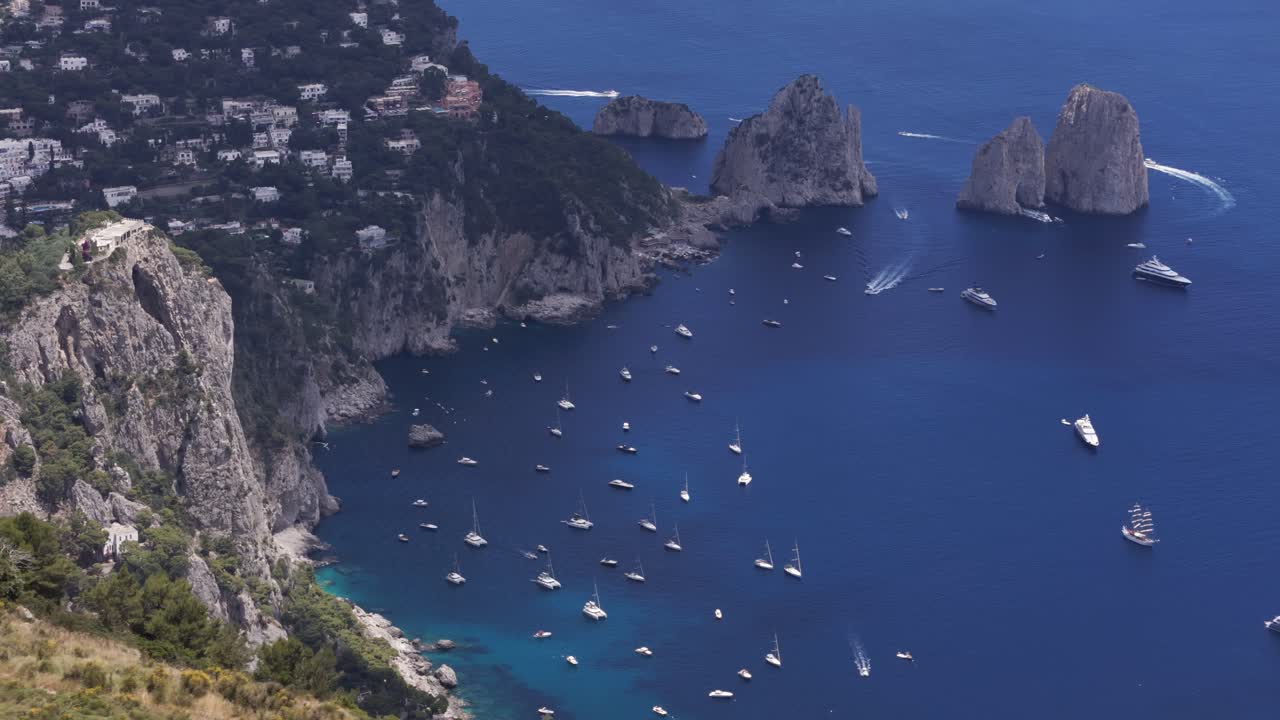 Aerial drone shot panning around the Faraglioni rocks with luxury boats sailing through the crystal-clear Mediterranean sea