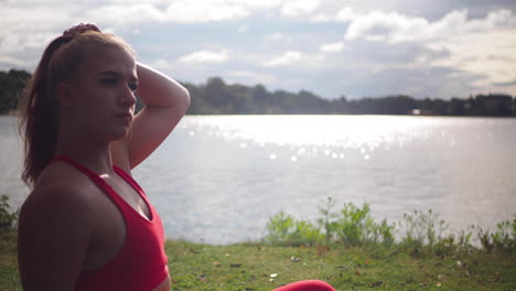 young woman in red sportswear sitting on the grass near lake and touching her long hair