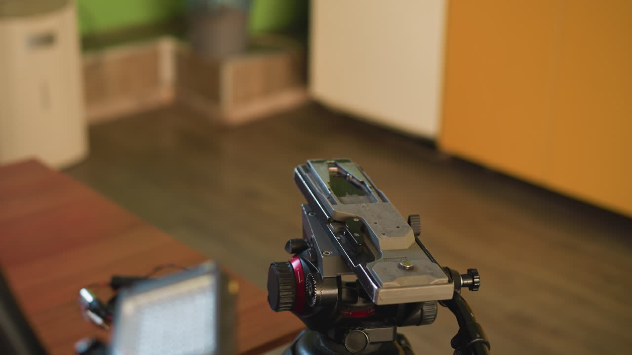Close-up shot of camera tripod head with focus on quick release plate. Microphone visible in foreground. Background shows home environment with green and orange elements