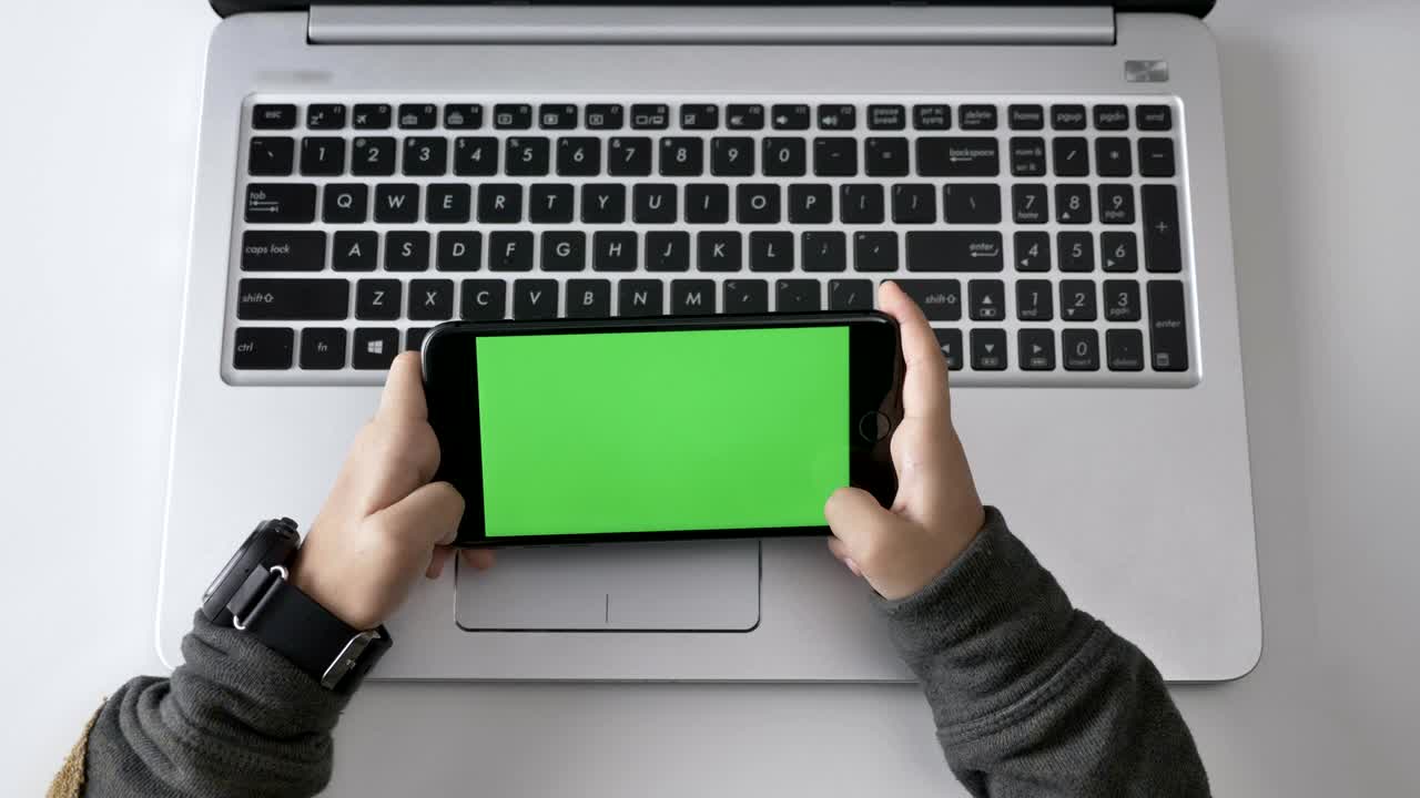 Children's hands hold a smartphone, Green screen, chromakey concept, laptop in the background. Top shot 60 fps