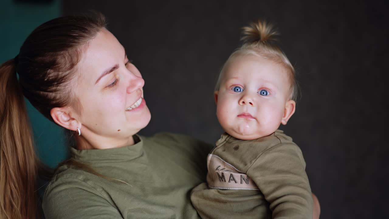 Caucasian mother is holding her beloved baby. Child looks ahead with focused and mom kisses her kid on the cheek.