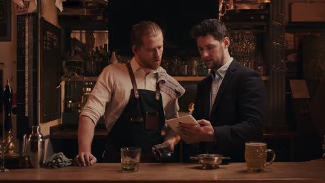 caucasian male bartender standing behind bar speaking to manager holding digital tablet