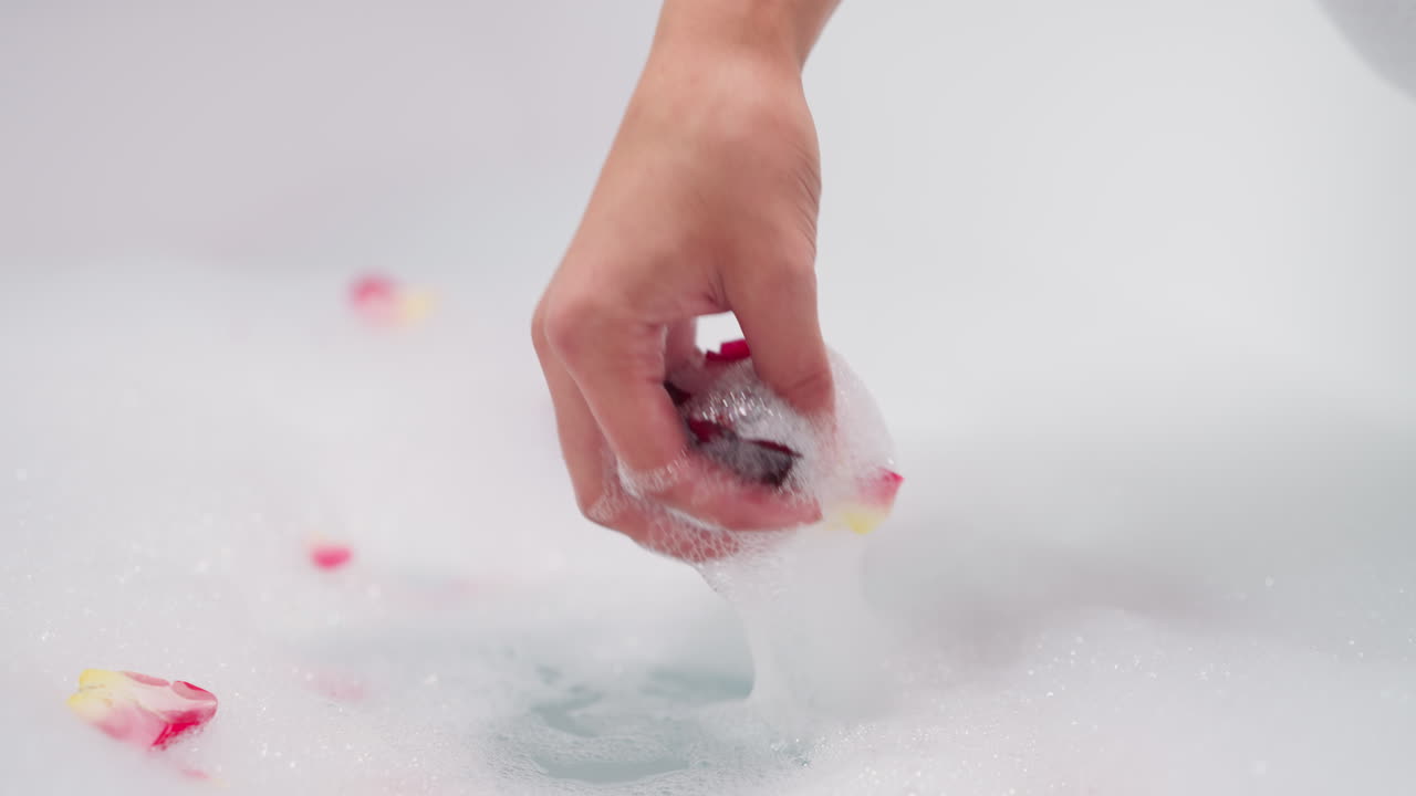 Lady grabs rose petals in foamy water closeup. Woman enjoys aromatic bubble bath featuring natural fresh flowers in bathtub. Spa salon experience