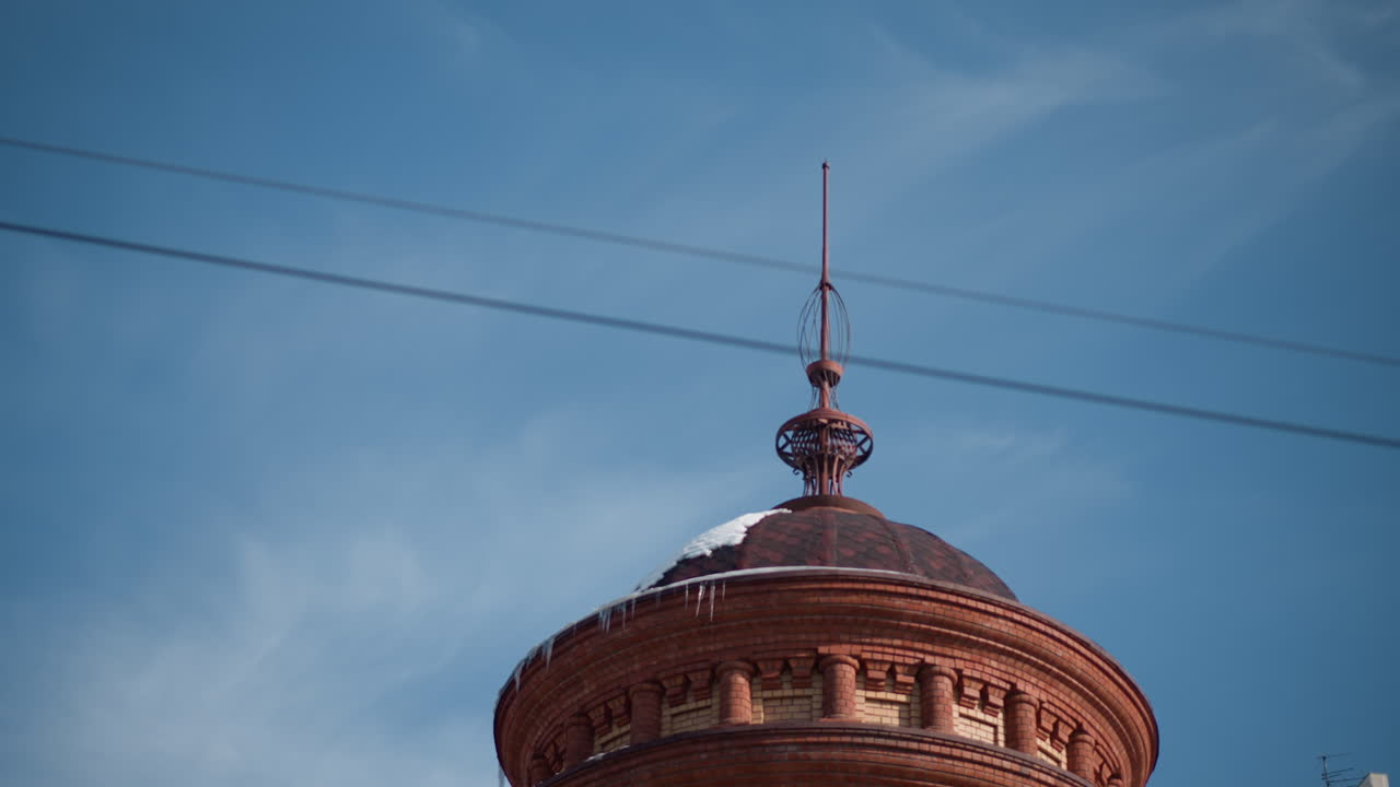 historic brick dome tower with ornate spire under blue winter sky, power lines crossing overhead, icicles hanging from roof cornice, urban landmark building detail against clouds, cold city scene