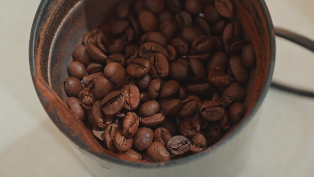 Primo piano di chicchi di caffè tostati nel macinacaffè, un accogliente rituale mattutino per gli amanti del caffè. Un cucchiaio versa i chicchi in un macinacaffè vintage di metallo, con ricche tonalità marroni, texture dettagliata e un aroma intenso, in un ritmo sottile.