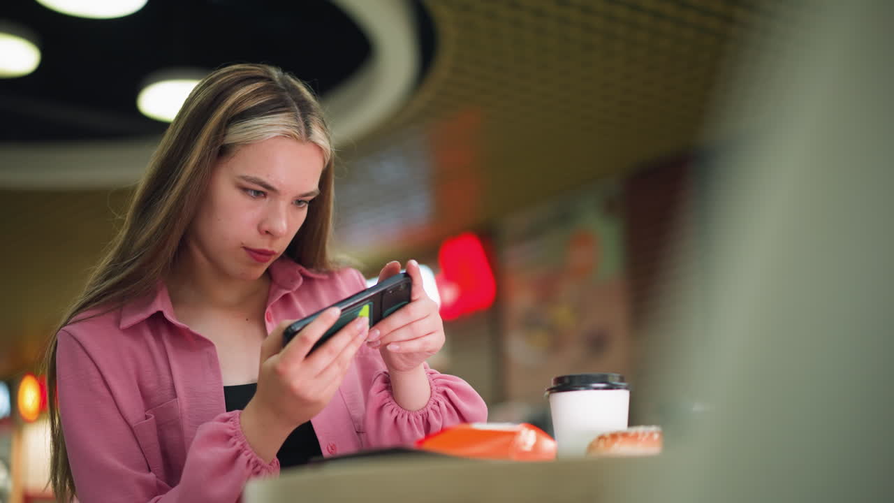 mujer de vestido rosa centrándose en su teléfono, posiblemente capturando una foto de comida delante de ella, taza de café y hamburguesa visible en la mesa, con un fondo borroso con iluminación ambiental