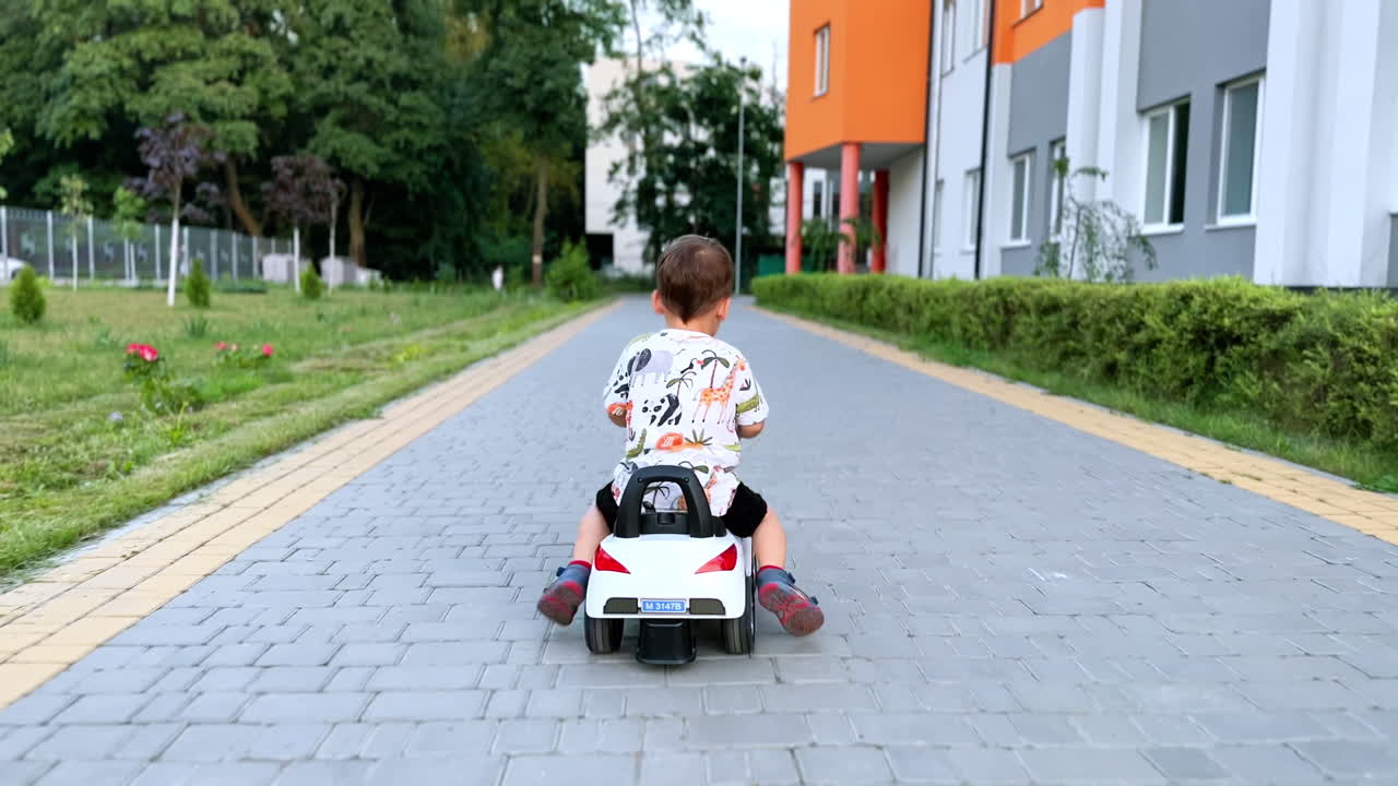 Rear view of a baby boy driving a toy car outdoors. Kid quickly moves his feet to push from the ground and ride.
