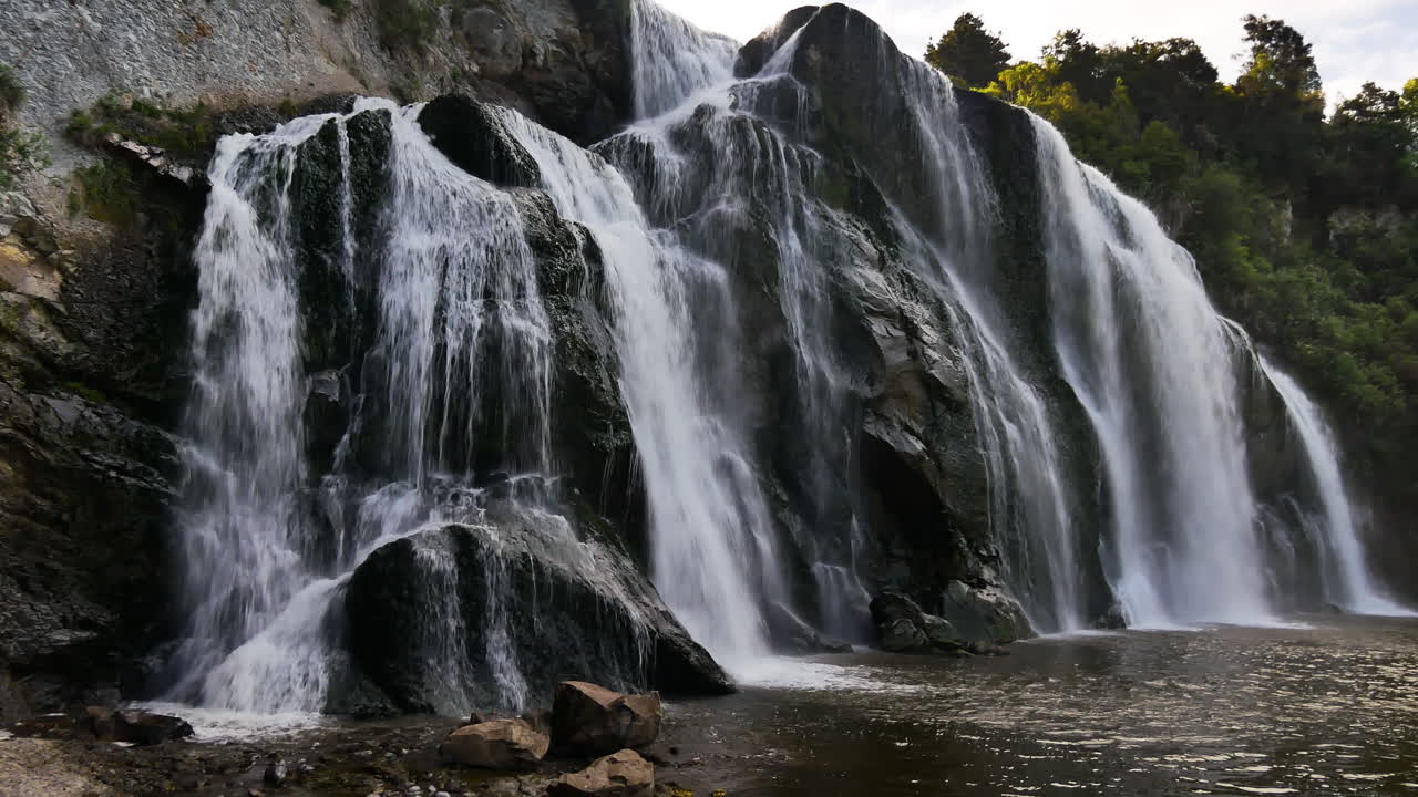 Panning shot of gigantic Waihi Waterfall flowing into idyllic lake in New Zealand, Manawatu-Wanganui