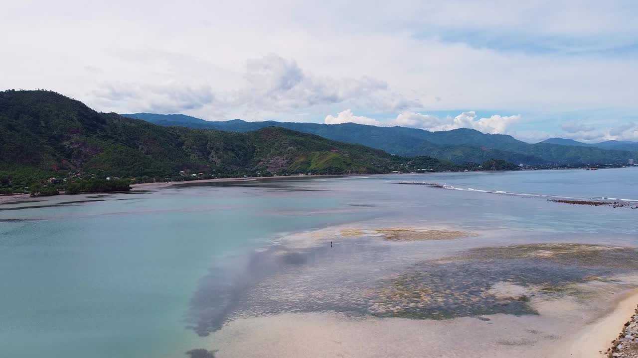 Aerial view rising over fisherman knee deep in ocean at low tide with rugged hills with trees in Dili, Timor-Leste, Southeast Asia